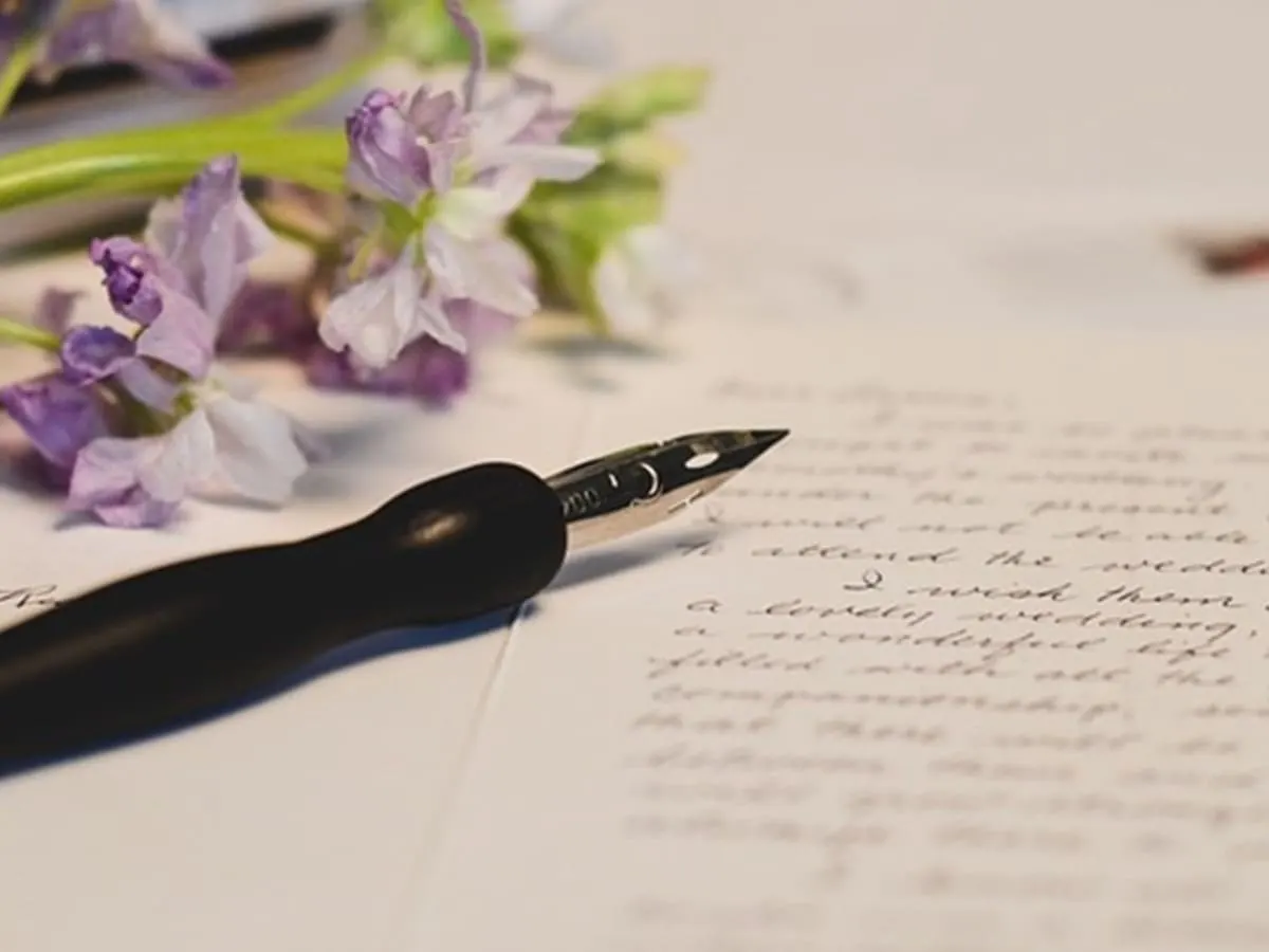 Une plume est posée sur une feuille à l'écriture manuscrite, à côté d'un bouquet de fleurs mauves.