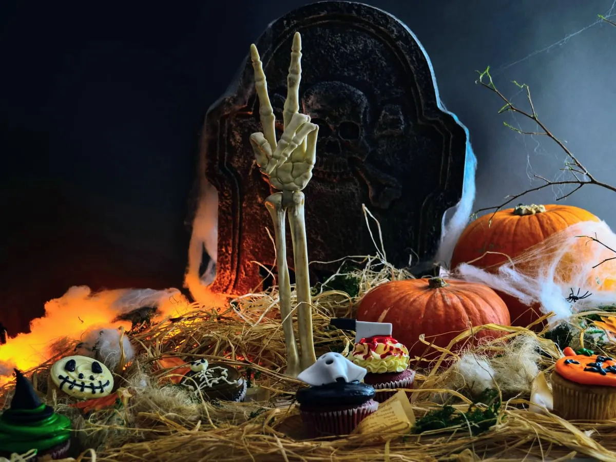 Une photo d'une main de squelette faisant le signe de la paix, émergeant d'un tas de paille et de fausses toiles d'araignée en avant d'une pierre tombale et des citrouilles. Des cupcakes sur le thème d'Halloween sont disposés dans la paille.