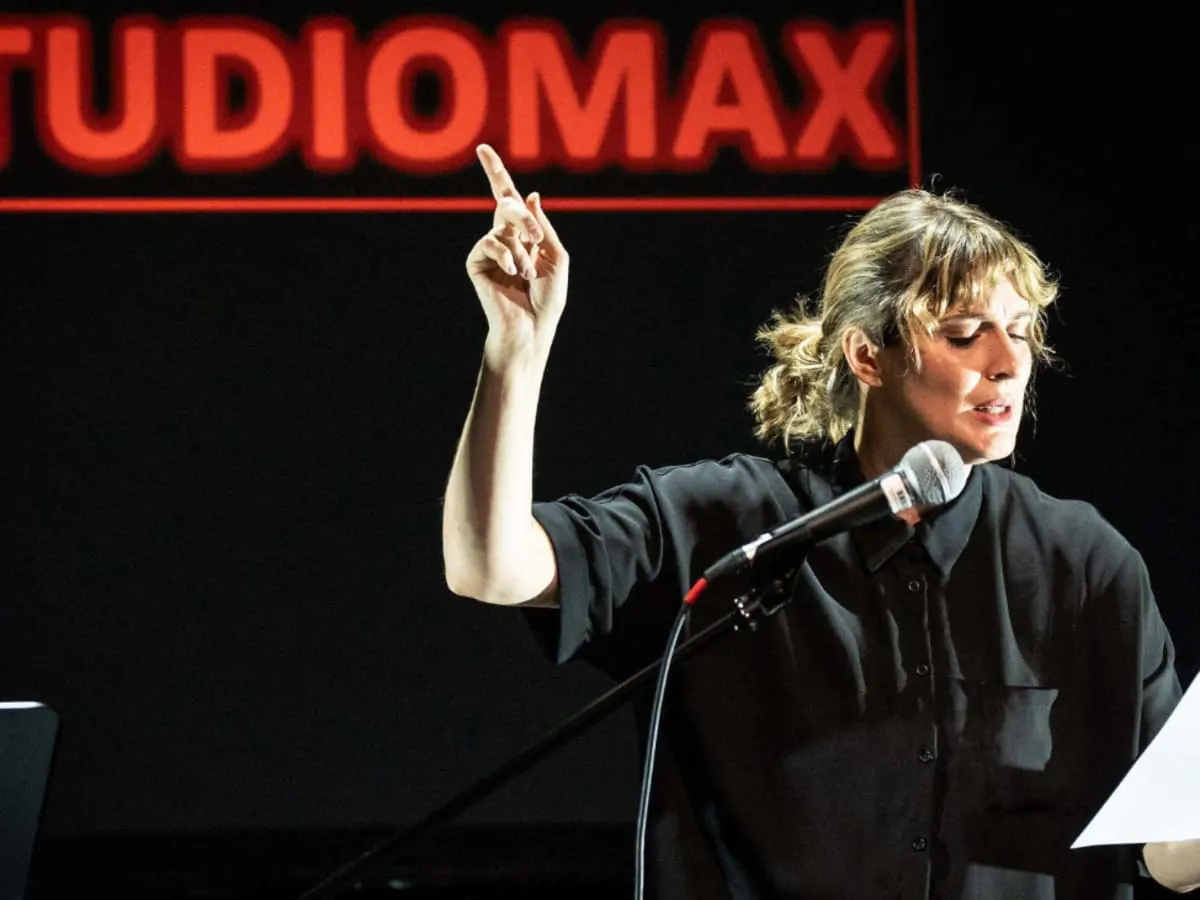 Une femme récite un texte au micro en levant l'index en l'air. En arrière d'elle se trouve une affiche où il est écrit: «STUDIOMAX».
