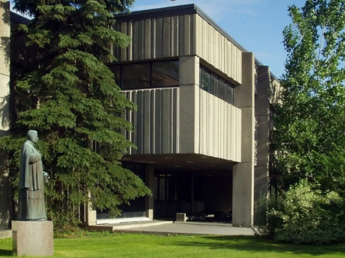 Bâtiment moderne entouré d’arbres, avec une statue sur socle au premier plan et une pelouse verdoyante devant l’entrée.