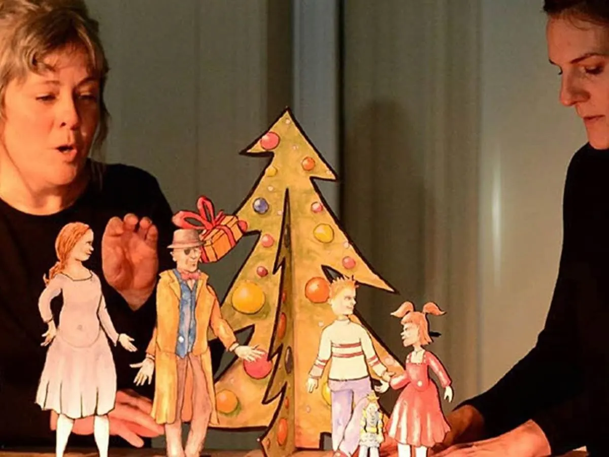 Deux personnes manipulent des marionnettes en papier autour d’un sapin sur une petite scène.