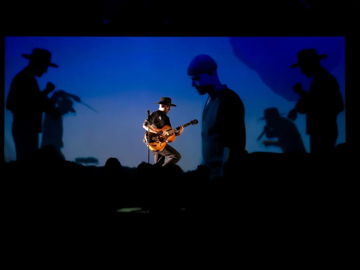 Un artiste portant un chapeau joue de la guitare sur scène avec son ombre en arrière-plan.