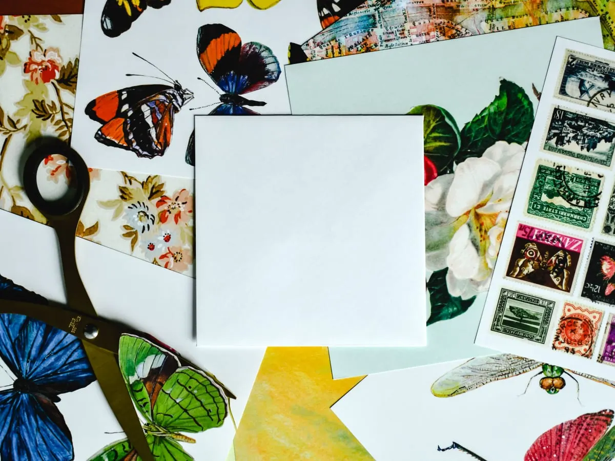 Photo d'une page blanche placé sur une pile de feuilles colorées ayant des dessins de papillons, fleurs et timbres.