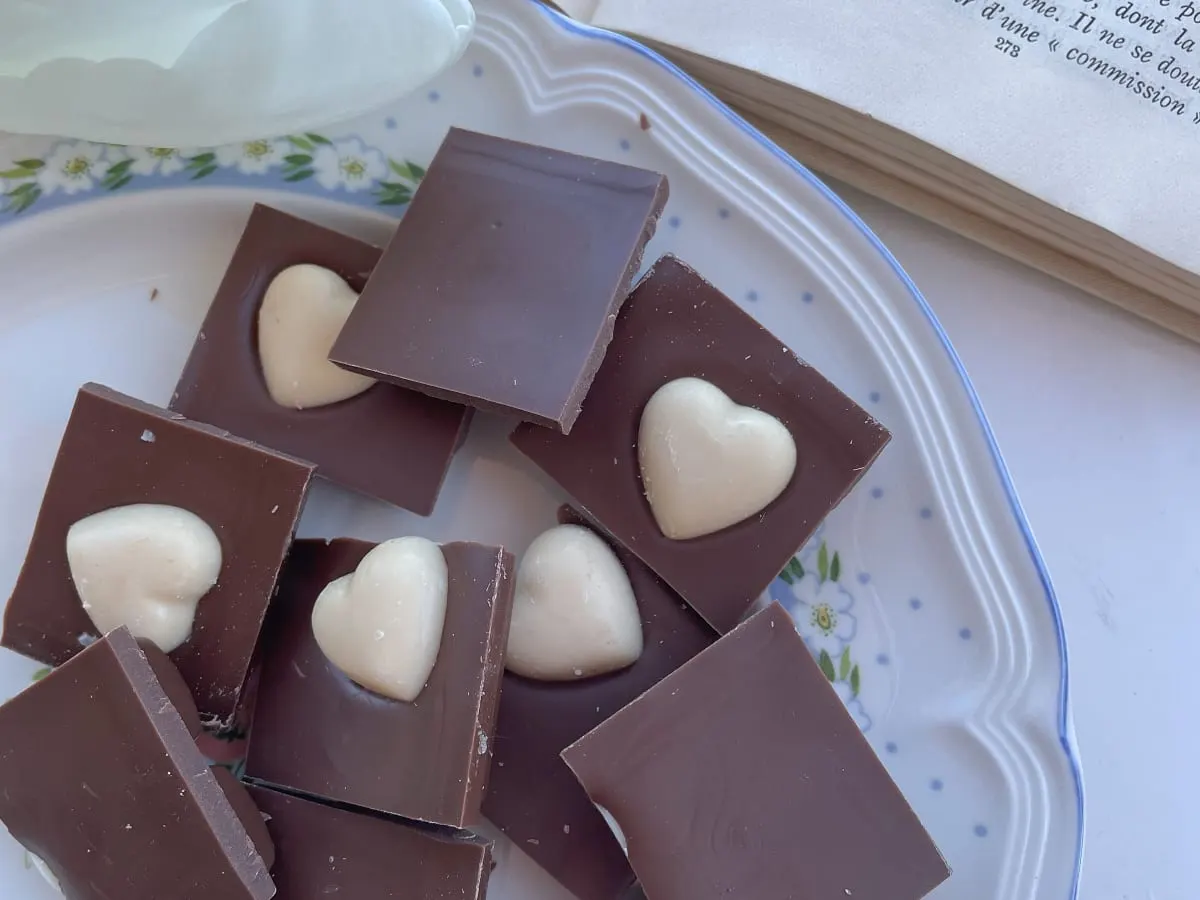 L'image présente une nature morte gourmande composée de morceaux de chocolat disposés sur une assiette décorative, accompagnés d'un livre ouvert.