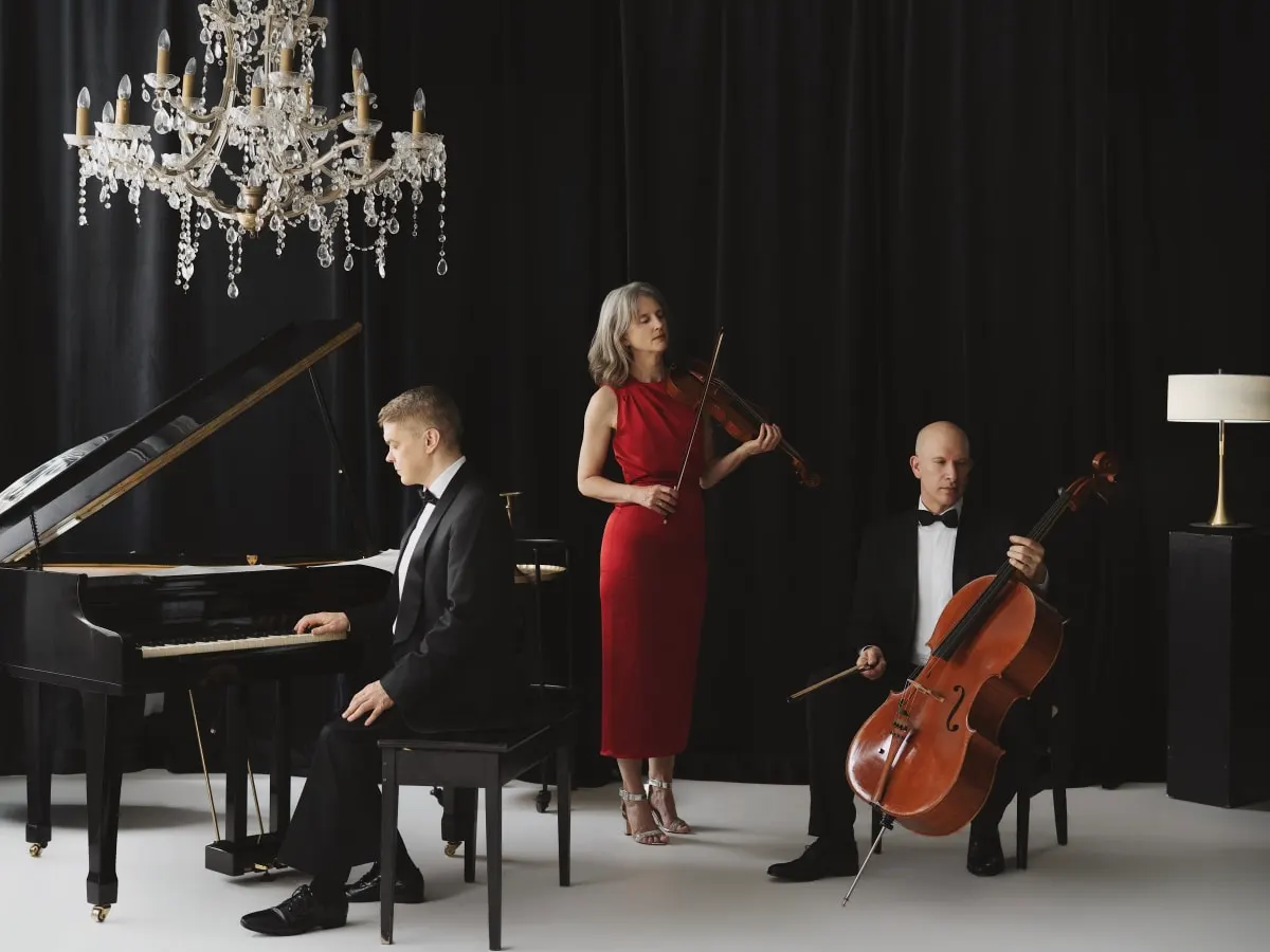 Trio de musique classique composé d’un pianiste, d’une violoniste et d’un violoncelliste sur une scène avec rideaux noirs, sous un lustre.