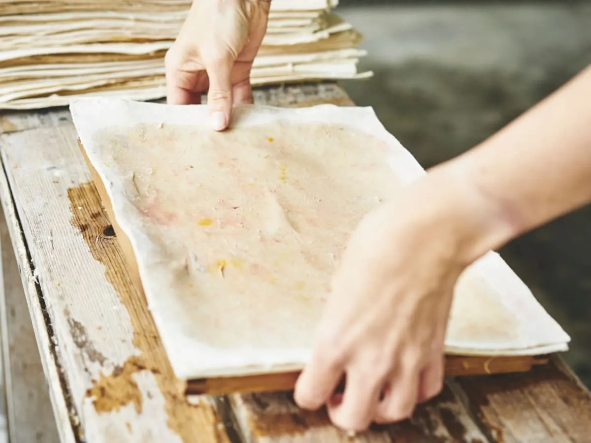 Des mains manipulent un tamis rectangulaire pour créer une feuille de papier artisanale.