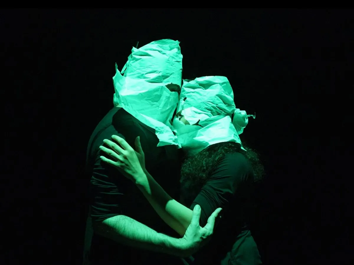 Couple de danseurs cagoulés et enlacés sur scène, avec arrière plan très sombre.
